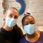 hannah and mosope looking at the camera, with masks on, a grey brick background with a blue logo.