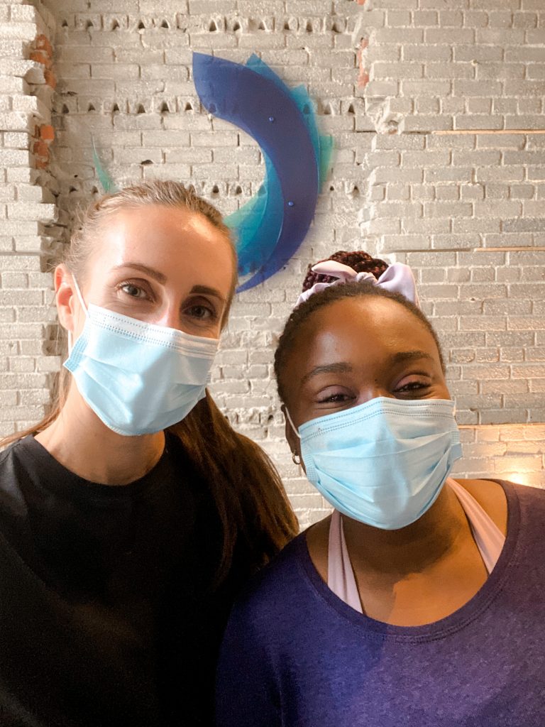 hannah and mosope looking at the camera, with masks on, a grey brick background with a blue logo.