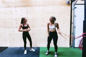 Hannah and Hannah in sports gear standing in a gym laughing, looking at each other.