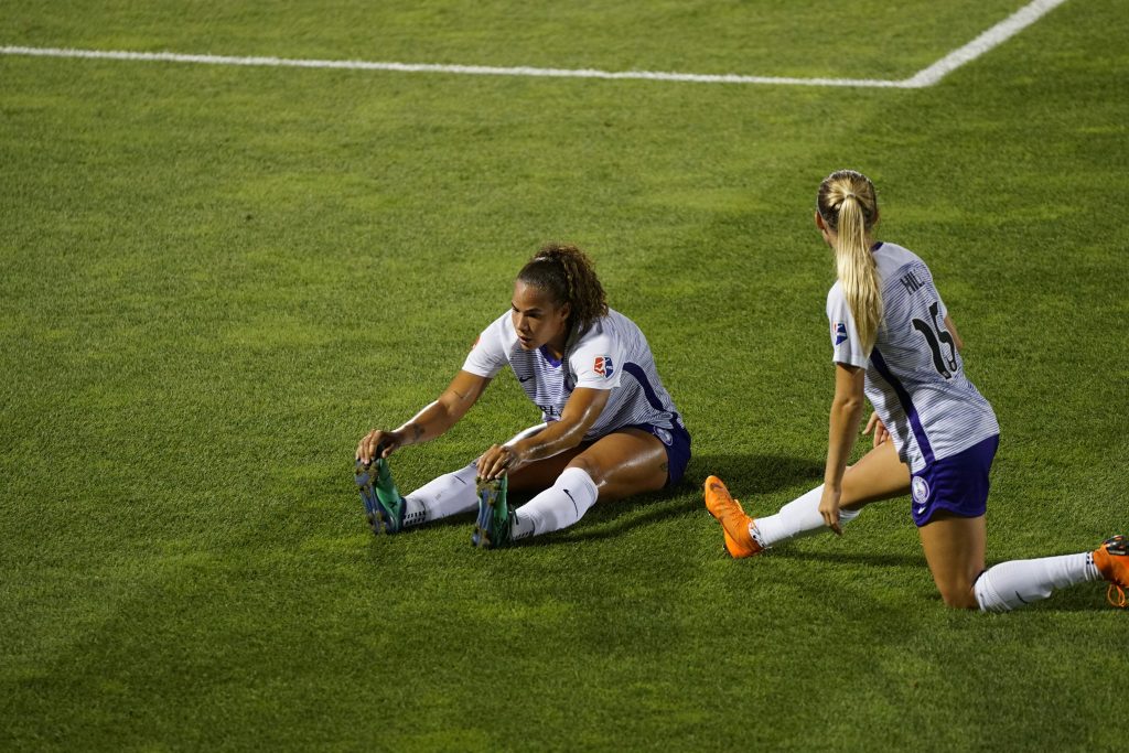 two female soccer players on the grass. one in an l-sit stretching and another in a half lunge stretching