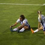 two female soccer players on the grass. one in an l-sit stretching and another in a half lunge stretching