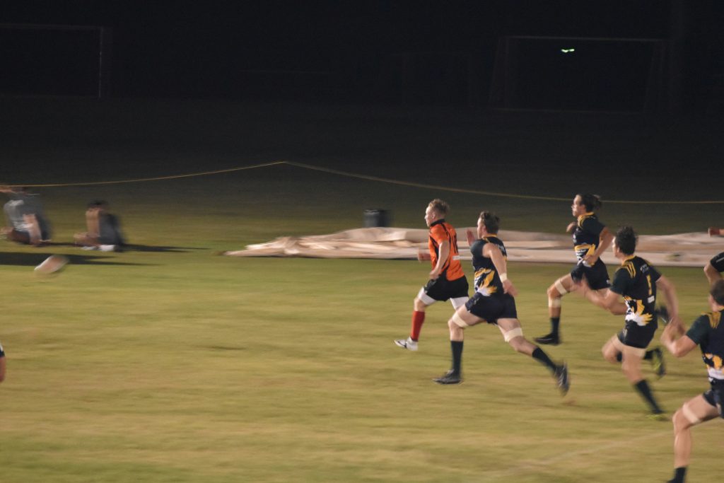 dark sky and green grass field with 6 men sprinting forwards on it in athletic team kits.