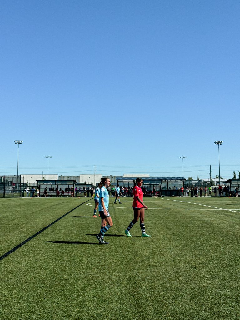 hannah on a grass field in a blue jersey playing soccer, standing next to the opposition player who is wearing red and black