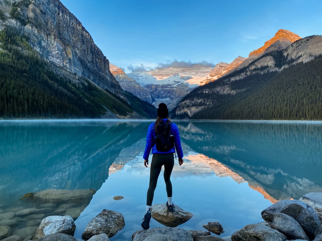 hannah standing on a rock in front of a blue lake with mountains in the background at sunrise