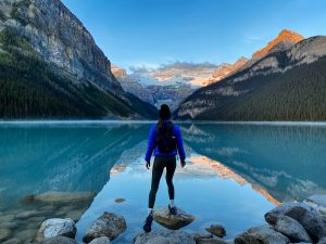 hannah standing on a rock in front of a blue lake with mountains in the background at sunrise